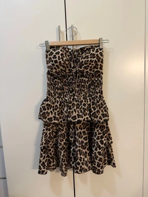 Leopardmönstrad kort klänning - Supersnygg kort klänning med leopardmönster och volanger. Klänningen är axelbandslös med smockad överdel och knyt fram. Perfekt för dig som vill sticka ut och älskar djurmönster!