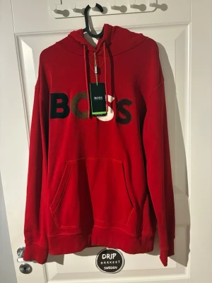 Röd hoodie från BOSS med logga - Röd hoodie från BOSS med stor broderad logga i svart, guld och vitt på bröstet. Klassisk känguruficka framtill, justerbar huva med snören och ribbade muddar. Perfekt för en chill och trendig stil.