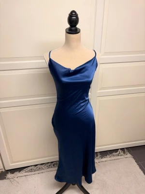 Marinblå långklänning i satin - Blå party-/cocktailklänning från Cider i stl S och normal i storleken. Tunn axelbandsmodell med korsade band i ryggen och lätt draperad ringning fram. Klänningen är helt ny med prislapp Vid frågor om mått eller annat är det bara att höra av sig💕