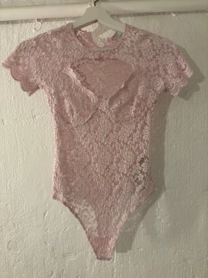Rosa spetsbody med cut out-detalj - Säljer en ljusrosa body i genombruten spets med korta ärmar och rund hals. Framsidan har en snygg cut out-detalj och inbyggd bystform. Materialet är stretchigt och följsamt, perfekt för att skapa en trendig look. Ingen synlig logga eller märke.