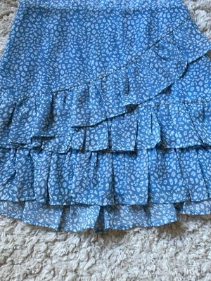 Blå volangkjol från Made by Chelsea - Supersöt blå kortkjol från Made by Chelsea med vitt leopardmönster och flera volanglager. Det är inga synliga defekter men på insidan av dragkedjan är det lite små trådar som sticker ut som inte syns utanpå. 
