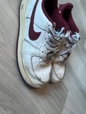 Nike Air Force 1 vita och vinröda - Klassiska Nike Air Force 1 sneakers i vitt läder med vinröda detaljer och sula. Skorna har perforerad tå, gråa skosnören och vinrött innerfoder. Den ikoniska swoosh-loggan syns tydligt på sidan. Perfekta för dig som gillar retrostil och streetwear.