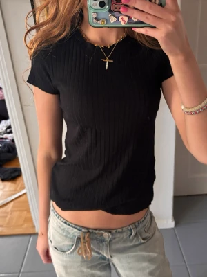 Svart ribbad kortärmad topp - En svart ribbad topp med korta ärmar och rund hals. Modellen är croppad och har en slim passform som sitter snyggt på kroppen. Perfekt till jeans eller kjol för en enkel och stilren look.