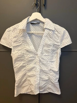 Vit prickig blus med kort ärm - Snygg vit blus med svarta små prickar och korta ärmar. Blusen har en klassisk krage, v-ringning och rynkade detaljer framtill som ger en cool och trendig look. Perfekt att matcha med jeans eller kjol för en fräsch outfit.