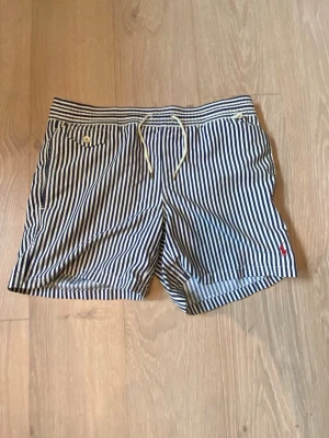 Ralph Lauren badshorts - Snygga shorts med marinblå och vita ränder, elastisk midja och dekorativa knappar på fickan. Perfekta för sommardagar och har en avslappnad passform. Tillverkade i mjukt bomullsmaterial för extra komfort. Liten gul missfärgning men knappt märkbart