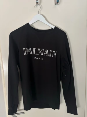 Svart sweatshirt från Balmain Paris - Stilren svart sweatshirt från Balmain Paris med vit logotyp på bröstet. Tröjan har rund halsringning, långa ärmar och ribbade muddar vid ärmslut och nederkant. Perfekt för dig som gillar exklusiva streetwear-plagg.