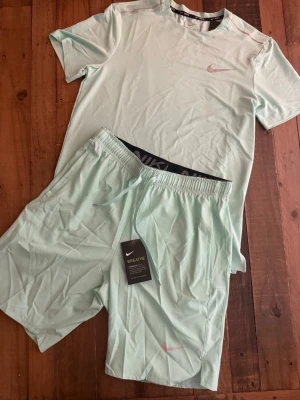 Nike Miller - Snyggt mintgrönt träningsset från Nike med t-shirt och shorts. Båda plaggen har diskret Nike-logga i rosa och är tillverkade i ett lätt, andningsbart material som passar perfekt för träning. Shortsen har dragsko i midjan och sidofickor.