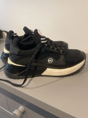 Svarta Michael Kors sneakers - Snygga svarta sneakers från Michael Kors med vita detaljer och grov sula. Skorna har MK-logga på sidan och bak, samt snörning framtill. Tillverkade i en mix av läder och textil för en modern look.