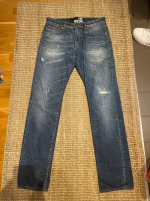 Love Moschino Jeans – Strl 32 | Distressed Denim - Säljer ett par Love Moschino-jeans i storlek 32. Snygg mellanblå tvätt med slitna detaljer som ger jeansen en modern och trendig look. Perfekta för dig som gillar designerplagg.