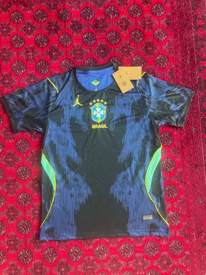 Brasil x Jordan matchtröja svart/blå - Snygg Brasilien matchtröja i samarbete med Jordan. Tröjan är svart och blå med gula och gröna detaljer, Brasil-logga och Jordan-logga på bröstet. Tillverkad i lätt och ventilerande funktionsmaterial, perfekt för fotboll eller träning.