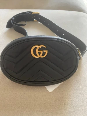 Svart magväska från Gucci - Snygg svart magväska från Gucci Marmonth i quiltat läder med GG-logga i guld framtill. Väskan har justerbart bälte och oval form, perfekt för att bära det viktigaste. Klassisk och trendig design med gulddetaljer. Äkta och med koden på insidan! Äktighetsintyg finns. Aldrig använd. 