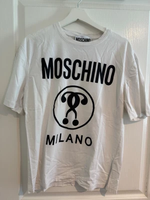 Vit Moschino Milano t-shirt - Säljer en vit t-shirt från Moschino med stor svart logga och Milano-tryck på framsidan. Klassisk rund hals och korta ärmar. Perfekt för dig som gillar streetwear och vill sticka ut med ett ikoniskt märke.