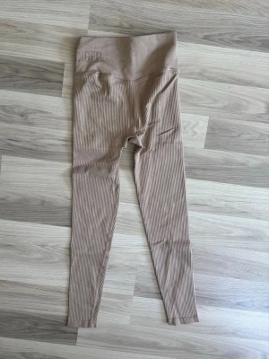 Beige ribbade tights från Stronger - Säljer ett par beige ribbade tights från Stronger. De har hög midja och är tillverkade i ett stretchigt material av 94% polyamid och 6% elastan. Perfekta för träning eller chill. Diskret logga vid midjan och snygg passform.