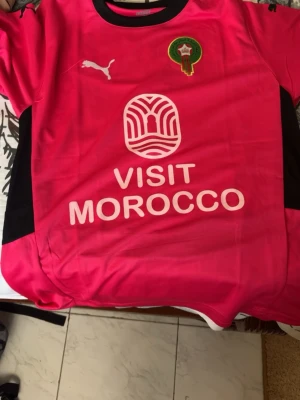 Marocko fotbollströja Puma röd - Snygg röd fotbollströja från Puma med Marockos landslagsemblem på bröstet och texten 'VISIT MOROCCO' framtill. Tröjan har svarta detaljer på sidorna och svart krage. Tillverkad i lätt och ventilerande material, perfekt för träning eller match.
