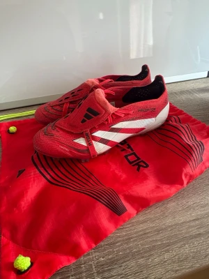 Adidas Predator röda fotbollsskor - Säljer ett par Adidas Predator fotbollsskor i rött med vita och svarta detaljer. Skorna har en strömlinjeformad design och är tillverkade i syntetmaterial för bra grepp och kontroll. Medföljer gör en matchande röd Predator-påse med svarta och gula detaljer. Skick 8,5/10. 