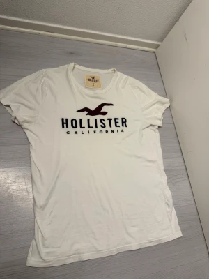 Vit Hollister t-shirt med logga - Vit t-shirt från Hollister med stor mörkröd logga och texten 'Hollister California' på bröstet. Klassisk rund halsringning och korta ärmar. Tillverkad i mjuk bomull för en skön känsla och avslappnad passform. Fraktar samma dag eller dagen efter :)
