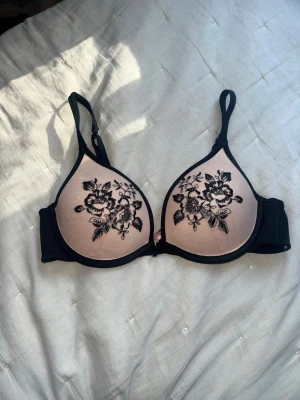 Hunkemöller BH, storlek 75B - Push-up bh i beige med svarta kanter och blommig spetsbrodyr på kuporna. Justerbara axelband och bygel för extra stöd. Kuporna är lätt vadderade och ger en fin form. 
