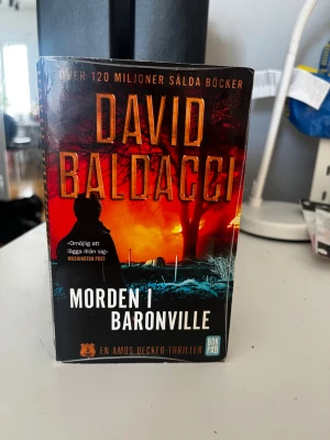 Morden i Baronville av David Baldacci - Spännande thrillerbok av David Baldacci, 'Morden i Baronville', i pocketformat. Omslaget har en mörk och dramatisk design med siluett och eldigt röd-orange bakgrund. Boken är en del av serien om FBI-utredaren Amos Decker och är på svenska.