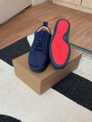 Mörkblå Christian louboutin sneakers med röd sula - Snygga mörkblå sneakers med ovandel i mocka och klassisk snörning. Skorna har en unik röd yttersula med grafiskt mönster och diskreta detaljer. Perfekta för dig som vill sticka ut med stilrena men ändå unika sneakers.