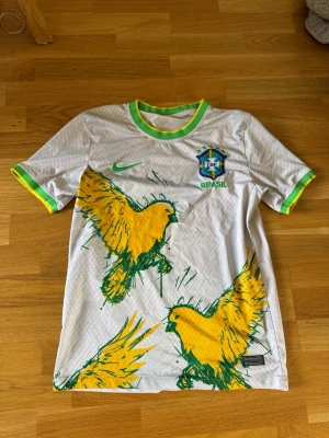 Vit Brasilien fågel fotbollströja Nike - Säljer en eftertraktad och populär vit Brasilien fotbollströja från Nike med gröna och gula detaljer. Tröjan har ett stort tryck med gula fåglar och det klassiska CBF-emblemet på bröstet. Rund hals med grönt och gult, samt korta ärmar. Perfekt för dig som älskar fotboll och vill sticka ut.