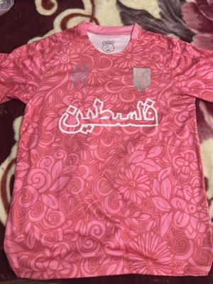 Rosa fotbollströja Palestina M - Säljer en rosa fotbollströja med blommigt mönster och vit text på arabiska över bröstet. Tröjan har rund halsringning och klubbmärke på vänster sida. Tillverkad i polyester, perfekt för träning eller match.