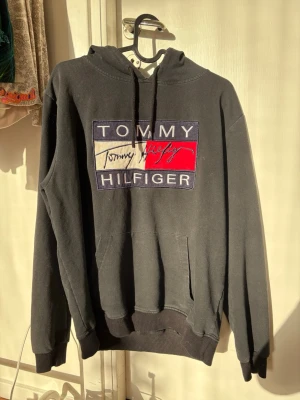 Svart hoodie Tommy Hilfiger - Säljer en svart hoodie från Tommy Hilfiger med stor broderad logga på bröstet i blått, vitt och rött. Hoodien har huva med dragsko och en klassisk magficka. Perfekt för dig som gillar streetwear och vill ha en stilren look.