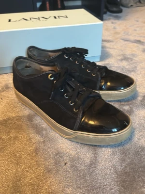 Svarta Lanvin sneakers med lackdetaljer - Snygga svarta sneakers från Lanvin med både mocka och glansiga lackdetaljer på tån och hälen. Skorna har svarta skosnören, guldfärgade öljetter och en beige sula som ger en clean kontrast. Perfekta för dig som gillar stilrena men ändå unika sneakers.