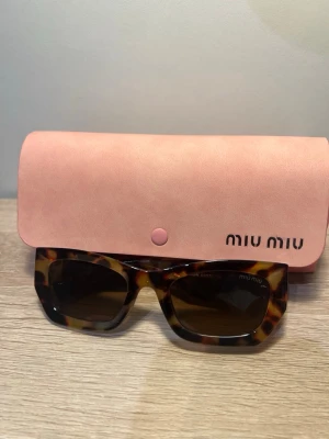 Turtleskalade solglasögon från Miu Miu - Snygga solglasögon från Miu Miu med fyrkantiga bågar i brun turtleskal och mörka linser. Kommer med ett rosa fodral med Miu Miu-logga. Perfekta för dig som vill ha en trendig accessoar med lyxig känsla.