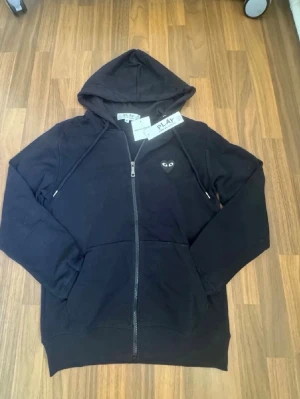 Comme des Garçons Play - Svart hoodie med dragkedja från Comme des Garçons Play. Har det ikoniska hjärta-med-ögon-trycket på bröstet och ett stort hjärta med ögon på ryggen. Mjuk bomull, huva med dragsko och fickor framtill. Perfekt för en streetwear-look.
