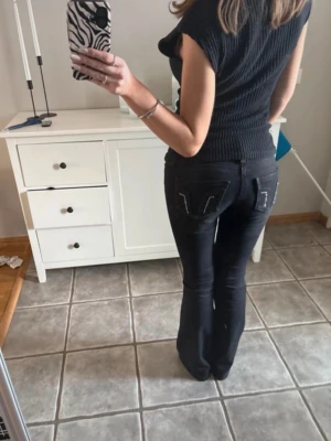 Svarta bootcut jeans med detaljer - Säljer ett par svarta bootcut jeans med snygga vita sömdetaljer på bakfickorna. Jeansen har klassisk femficksdesign och är tillverkade i denim med stretch för skön passform. Perfekta för dig som gillar en trendig och bekväm stil. Säljer då de är för små, paljetterna på bakfickorna har ramlat av i tvätten.
