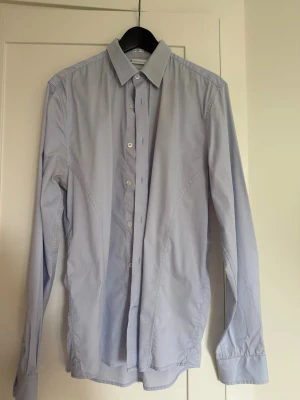Ljusblå skjorta från These Glory Days - Snygg ljusblå skjorta från These Glory Days i stretchig bomull. Skjortan har slim fit-passform, klassisk krage och knäppning framtill. Perfekt för dig som gillar stilren och enkel design.