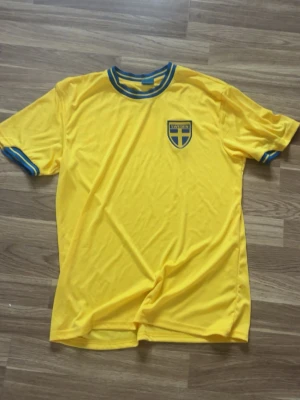 Sverige fotbollströja - Gul fotbollströja med blå detaljer och Sveriges landslagsemblem på bröstet. På ryggen står det 'SVERIGE' i stora vita bokstäver och en liten svensk flagga ovanför. Perfekt för att heja på landslaget eller spela match.