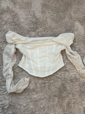 Vit offshoulder korsettopp - Trendig vit korsettopp med offshoulder och långa puffärmar. Toppen har en croppad passform och markerade sömmar framtill för en snygg siluett. Gåva vid köp!🎁