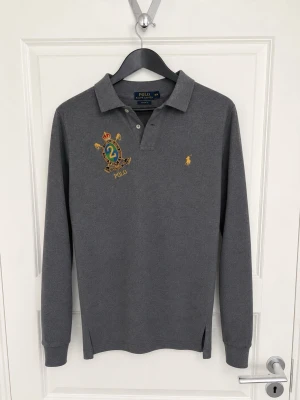 Grå långärmad piké från Ralph Lauren - Snygg grå långärmad pikétröja från Polo Ralph Lauren med broderad logga och emblem på bröstet. Tröjan har klassisk krage, knappar vid halsen och ribbade muddar. Perfekt för en stilren och sportig look.
