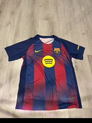 Barcelona matchtröja Nike blå/röd - Snygg FC Barcelona matchtröja från Nike i blått och rött med gula detaljer. Tröjan har klubbmärke på bröstet, gul Nike-logga och Spotify som sponsor framtill. Tillverkad i lätt och ventilerande material, perfekt för fotboll eller träning.
