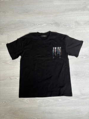 Svart AMIRI t-shirt med tryck - Säljer en svart t-shirt från AMIRI med färgstarkt tryck på bröstet där bokstäverna har droppande effekt i blått, rött och vitt. Klassisk rund hals och korta ärmar. Perfekt för dig som gillar streetwear och vill sticka ut lite extra. Stor i storlek!