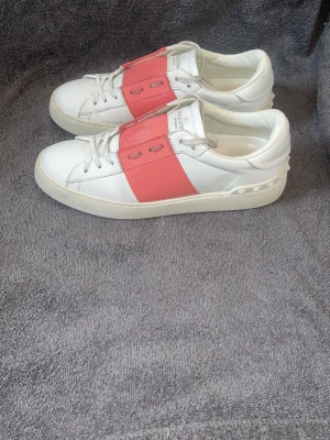 Valentino open - Snygga vita sneakers från Valentino med bred rosa panel över snörningen och klassisk logga på plösen. Skorna har en chunky sula och är tillverkade i slätt läder med coola nitar bak på hälen. Perfekta för dig som vill sticka ut med stilrena detaljer.