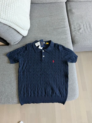 Marinblå kabelstickad polotopp Polo Ralph Lauren - Snygg marinblå kabelstickad polotopp från Polo Ralph Lauren med korta ärmar och klassisk krage. Toppen har två knappar framtill och den ikoniska röda loggan broderad på bröstet. Perfekt för en stilren och avslappnad look. Storlek M