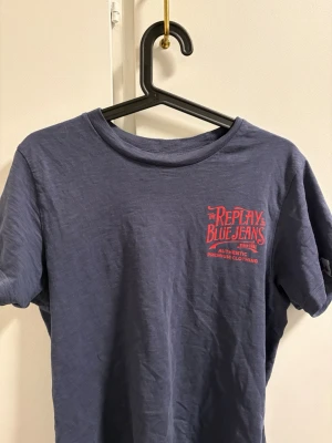 Blå Replay Blue Jeans t-shirt - Snygg blå t-shirt från Replay Blue Jeans med röd logga och text både fram och bak. Klassisk rund hals och korta ärmar. Tillverkad i mjuk bomull som känns skön mot huden. Perfekt för dig som gillar streetwear och vill ha en cool vardagslook.