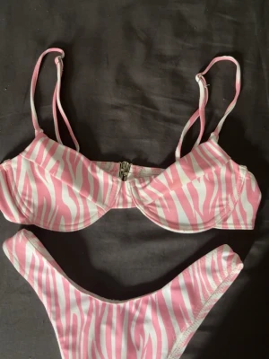 Rosa och vit zebramönstrad bikini - Säljer en snygg bikini med rosa och vitt zebramönster. Överdelen har justerbara axelband och en metallknäppning framtill. Bikinin har en modern, tight passform och är tillverkad i ett stretchigt syntetmaterial som passar perfekt till sommaren.