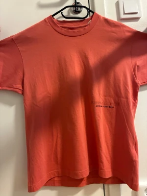 Korallröd Adidas t-shirt - Snygg korallröd t-shirt från Adidas med trycket 'FUSSBALLLIEBE' och 'ADIDAS FOOTBALL' på bröstet. Klassisk passform med rund halsringning och Adidas-logga i samma färg på ryggen. Tillverkad i mjuk bomull för skön känsla.