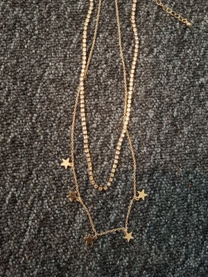 Guldigt halsband med stjärnor - Snyggt dubbelt halsband i guldfärg med små stjärnhängen och pärlliknande detaljer. Kedjan är tunn och smidig, perfekt för att ge din outfit lite extra bling. Passar dig som gillar trendiga accessoarer med en lekfull touch.
