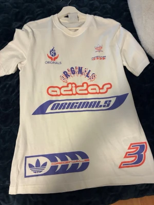 Vit Adidas Originals t-shirt - Säljer en vit Adidas Originals t-shirt med flera tryck i rött och blått både fram och bak. T-shirten har korta ärmar, rund hals och är gjord i mjuk bomull. Flera coola loggor och grafiska detaljer ger en unik streetwear-look.
