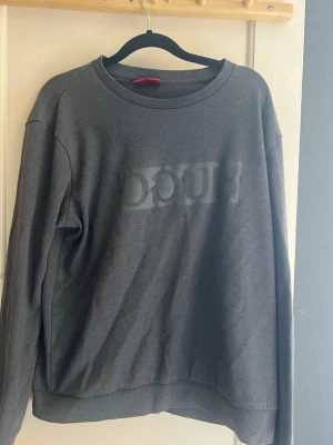 Mörkgrå sweatshirt från Hugo - Mörkgrå sweatshirt från Hugo med diskret logga framtill i samma ton. Klassisk rund halsringning och ribbade muddar vid ärmslut och nederkant. Perfekt för en avslappnad och stilren look. Storlek:L men sitter som small. Passar om du är runt 175 cm