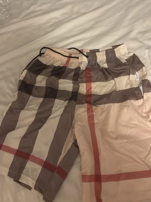  shorts - Säljer ett par rutiga shorts med bred resår i midjan och snörning. Shortsen har ett klassiskt beige, brunt, vitt och rött mönster och är tillverkade i ett lätt syntetmaterial. Perfekta för sommaren och riktigt sköna att bära.