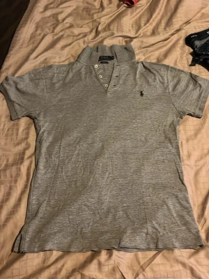 Grå pikétröja från Polo Ralph Lauren - grå pikétröja från Polo Ralph Lauren med korta ärmar, krage och knappar framtill. Tröjan har den ikoniska lilla broderade loggan på bröstet och är tillverkad i mjuk bomull. Perfekt till jeans eller shorts för en avslappnad look. Denna är perfekt inför sommaren och det är ett riktigt klipp för bara 349kr och nypris ligger på 1095kr säljer endast för 349kr eftersom den inte kommer till användning och den har även ett väldigt litet hål längst ner på tröjan som knappast syns därför sänks priset 