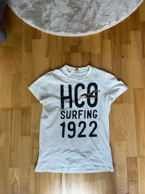 Vit HCO t-shirt med tryck - Vit t-shirt från Hollister med stort mörkblått tryck 'HCO Surfing 1922' och en röd fågel på bröstet. Klassisk rund hals och korta ärmar. Perfekt för dig som gillar surfstil och avslappnad look.