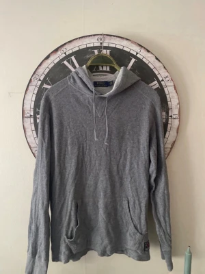 Grå hoodie från Polo Ralph Lauren - Snygg grå hoodie från Polo Ralph Lauren med huva och dragsnören. Tillverkad i mjukt material och har två fickor framtill. Perfekt för en avslappnad och stilren look.