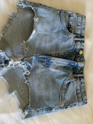 Ljusa jeansshorts med fransig kant - Säljer ett par ljusblå jeansshorts med fransiga kanter och slitna detaljer. Shortsen har klassisk femficksdesign, bälteshällor och dragkedja framtill. Perfekta för sommardagar och ger en avslappnad vibe.