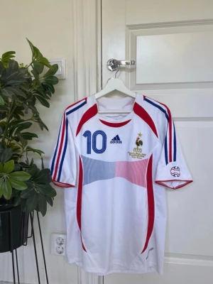 Frankrike Zidane tröja Adidas - Säljer en vit fotbollströja från Adidas med Frankrikes landslagsmärke, nummer 10 och Zidane på ryggen. Tröjan har blå och röda detaljer, korta ärmar och klassiska Adidas-ränder på axlarna. Perfekt för dig som älskar fotboll och vill ha en ikonisk supportertröja.